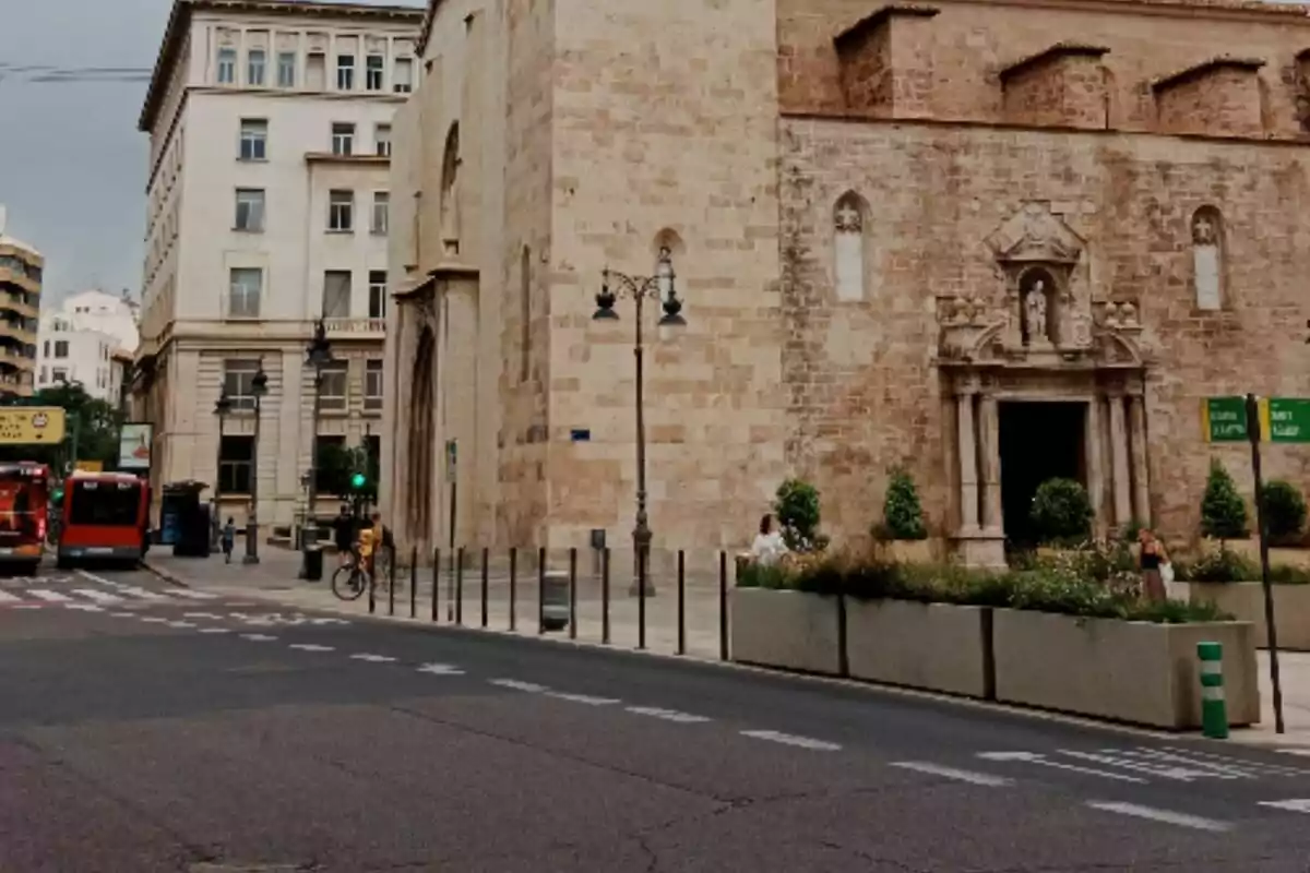 Calle urbana con una iglesia de piedra antigua, personas caminando, autobuses rojos y edificios altos al fondo ubicada en Valencia. Calle urbana con una iglesia de piedra antigua, personas caminando, autobuses rojos y edificios altos al fondo ubicada en Valencia.