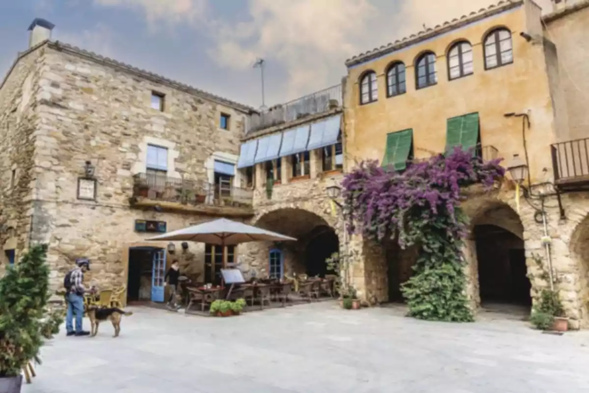 Plaça pintoresca amb edificis de pedra, terrassa de cafeteria, plantes i una persona passejant un gos Plaça pintoresca amb edificis de pedra, terrassa de cafeteria, plantes i una persona passejant un gos