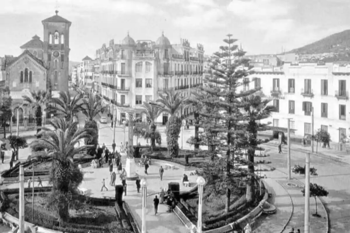 Fotografia antiga en blanc i negre d’una plaça amb jardins, palmeres, un gran arbre al centre, persones passejant i edificis històrics al voltant. Fotografia antiga en blanc i negre d’una plaça amb jardins, palmeres, un gran arbre al centre, persones passejant i edificis històrics al voltant.