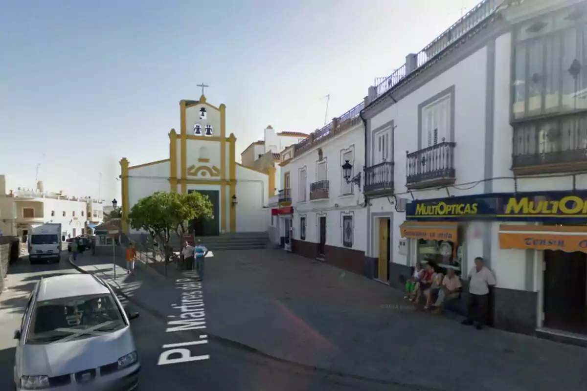 Vista de una plaza de Sevilla con una iglesia de fachada blanca y detalles amarillos al fondo, personas sentadas y de pie junto a una óptica y varios edificios residenciales a la derecha, un coche y un camión circulan por la calle. Vista de una plaza de Sevilla con una iglesia de fachada blanca y detalles amarillos al fondo, personas sentadas y de pie junto a una óptica y varios edificios residenciales a la derecha, un coche y un camión circulan por la calle.