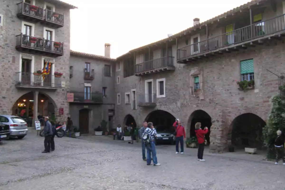 Persones passejant i fent fotos en una plaça envoltada d’edificis antics de pedra amb balcons i arcs Persones passejant i fent fotos en una plaça envoltada d’edificis antics de pedra amb balcons i arcs