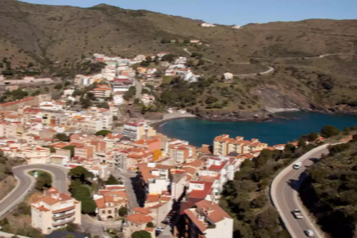 Vista aèria d’un poble costaner amb cases de teulades vermelles, una badia d’aigües blaves i muntanyes al fons Vista aèria d’un poble costaner amb cases de teulades vermelles, una badia d’aigües blaves i muntanyes al fons