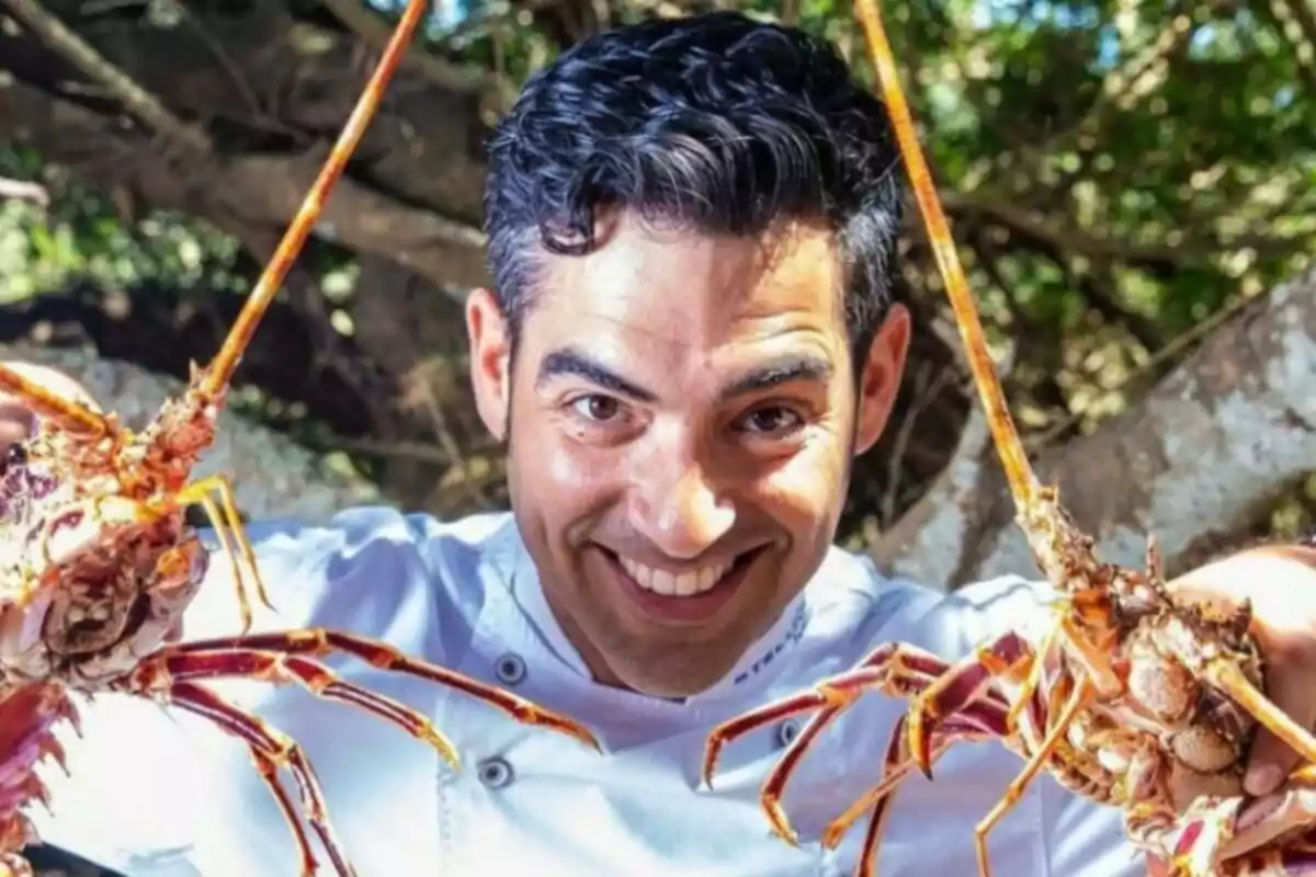 Andreu Genestra somrient amb cabells foscos i bata de xef sostenint dues llagostes grosses en un entorn natural Andreu Genestra somrient amb cabells foscos i bata de xef sostenint dues llagostes grosses en un entorn natural