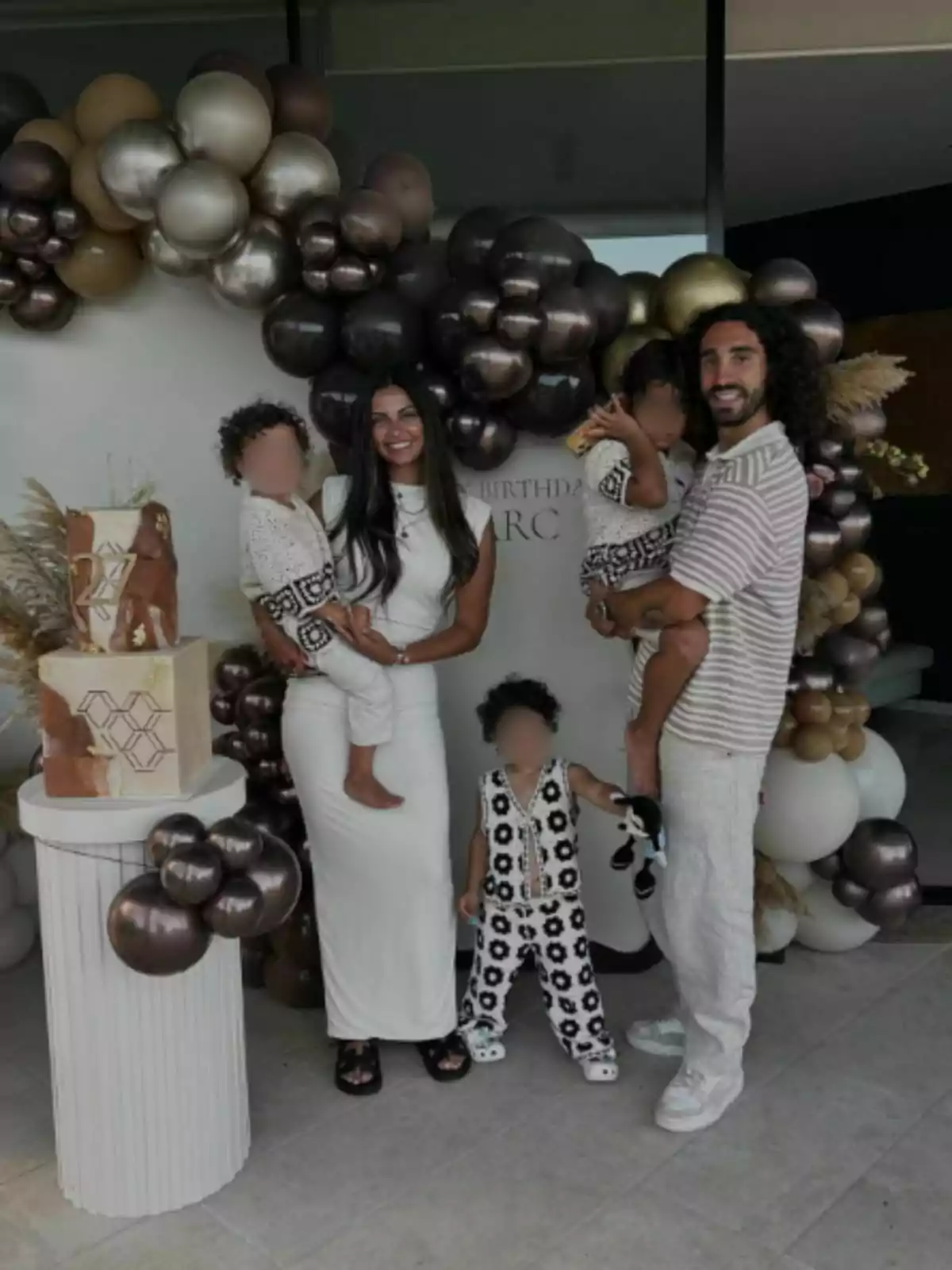 Familia posando sonriente frente a una decoración de globos en tonos dorados y marrones junto a un pastel de cumpleaños Familia posando sonriente frente a una decoración de globos en tonos dorados y marrones junto a un pastel de cumpleaños