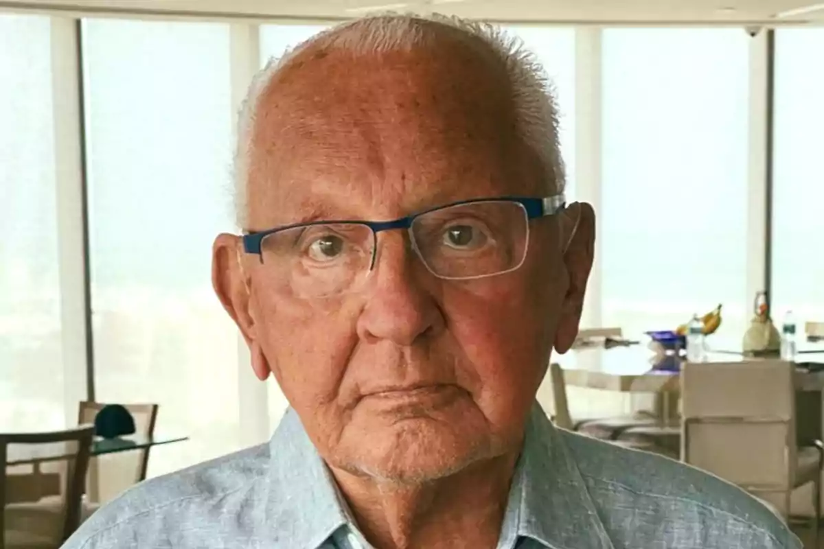 Older man with glasses and gray hair looking at the camera in a bright room with large windows in the background Older man with glasses and gray hair looking at the camera in a bright room with large windows in the background