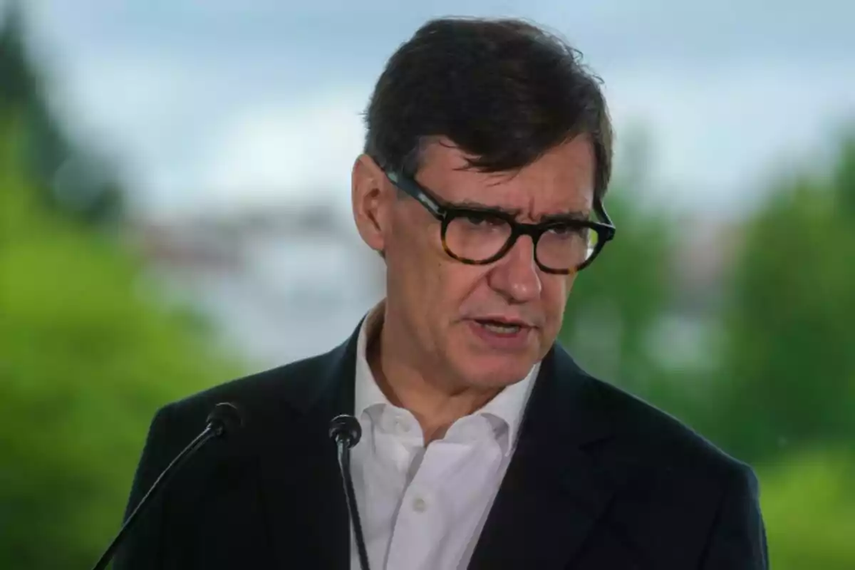 Hombre de cabello corto y gafas hablando frente a dos micrófonos con fondo de árboles desenfocados Hombre de cabello corto y gafas hablando frente a dos micrófonos con fondo de árboles desenfocados