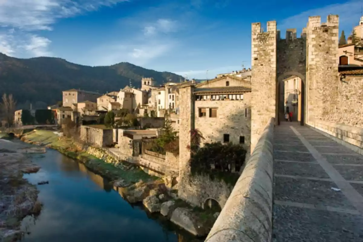 Pont medieval de pedra sobre un riu amb un poble antic i muntanyes al fons sota un cel blau Pont medieval de pedra sobre un riu amb un poble antic i muntanyes al fons sota un cel blau