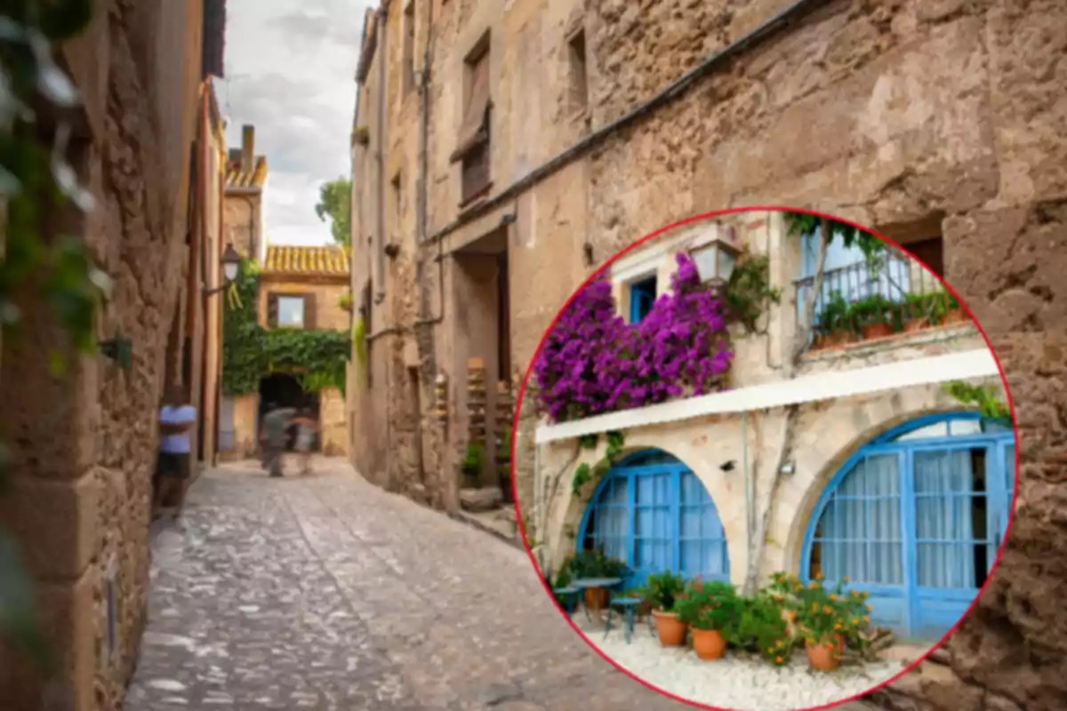 Callejón empedrado con casas de piedra y una ventana circular que muestra una fachada con puertas azules y flores moradas en el balcón