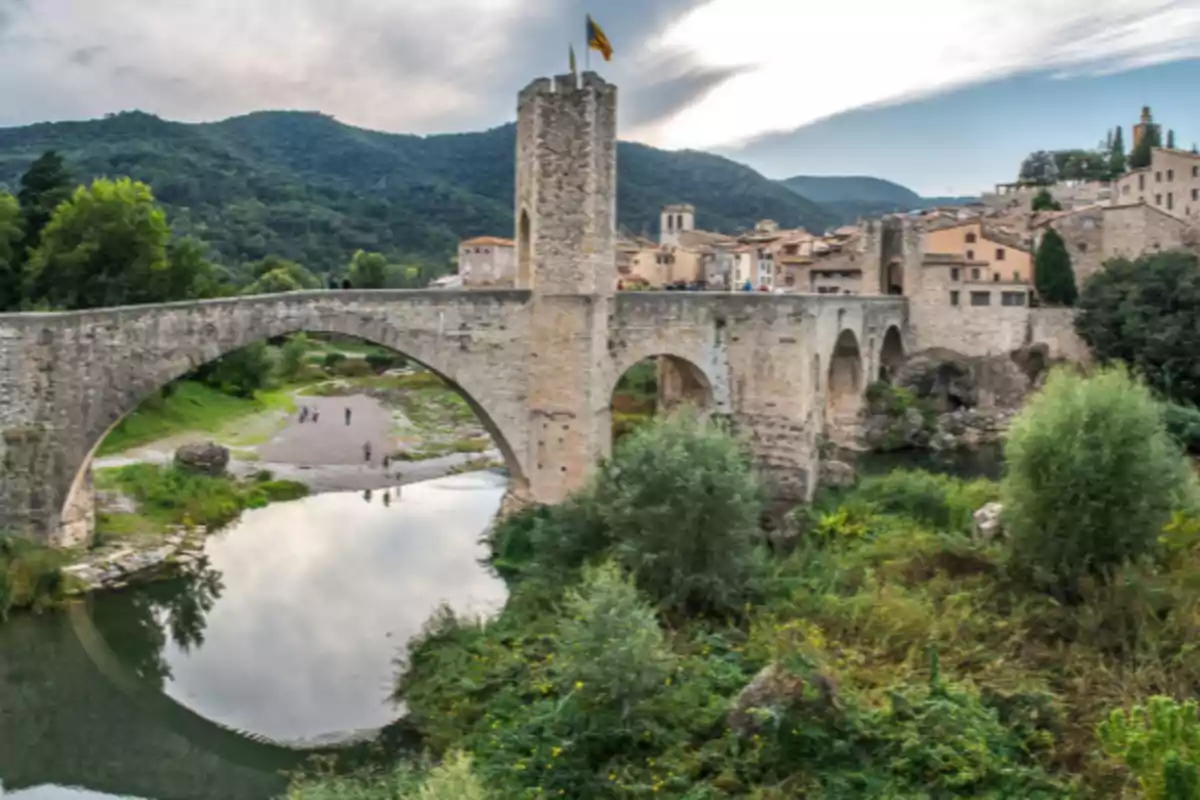 Pont medieval de pedra sobre un riu envoltat de vegetació, amb un poble antic al fons i muntanyes sota un cel ennuvolat Pont medieval de pedra sobre un riu envoltat de vegetació, amb un poble antic al fons i muntanyes sota un cel ennuvolat