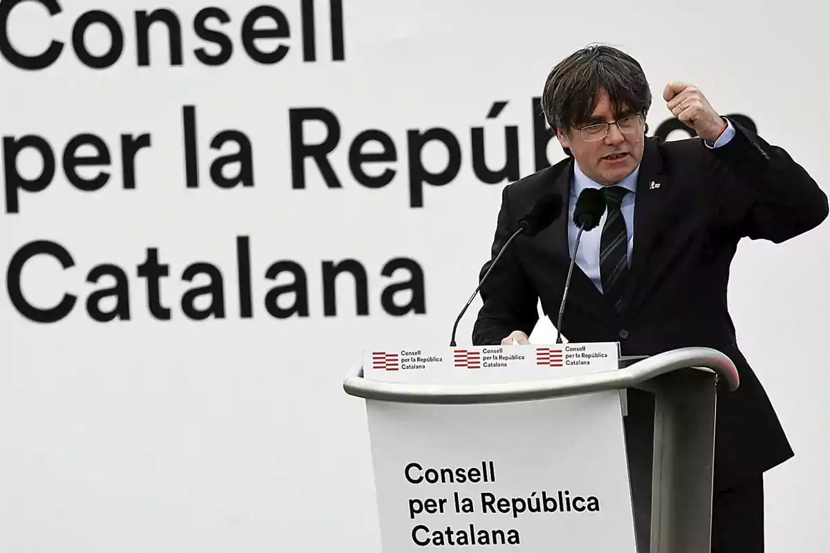 Hombre con gafas y traje hablando en un atril durante un evento del Consell per la República Catalana con el fondo de un cartel con el mismo texto