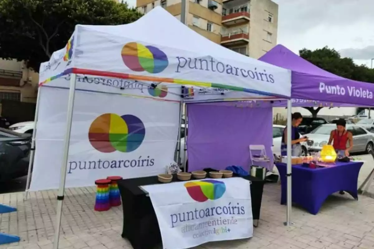 Dos carpas en la calle, una blanca con el logo y nombre "puntoarcoíris" y otra morada con el nombre "Punto Violeta", con mesas y personas organizando materiales sobre ellas