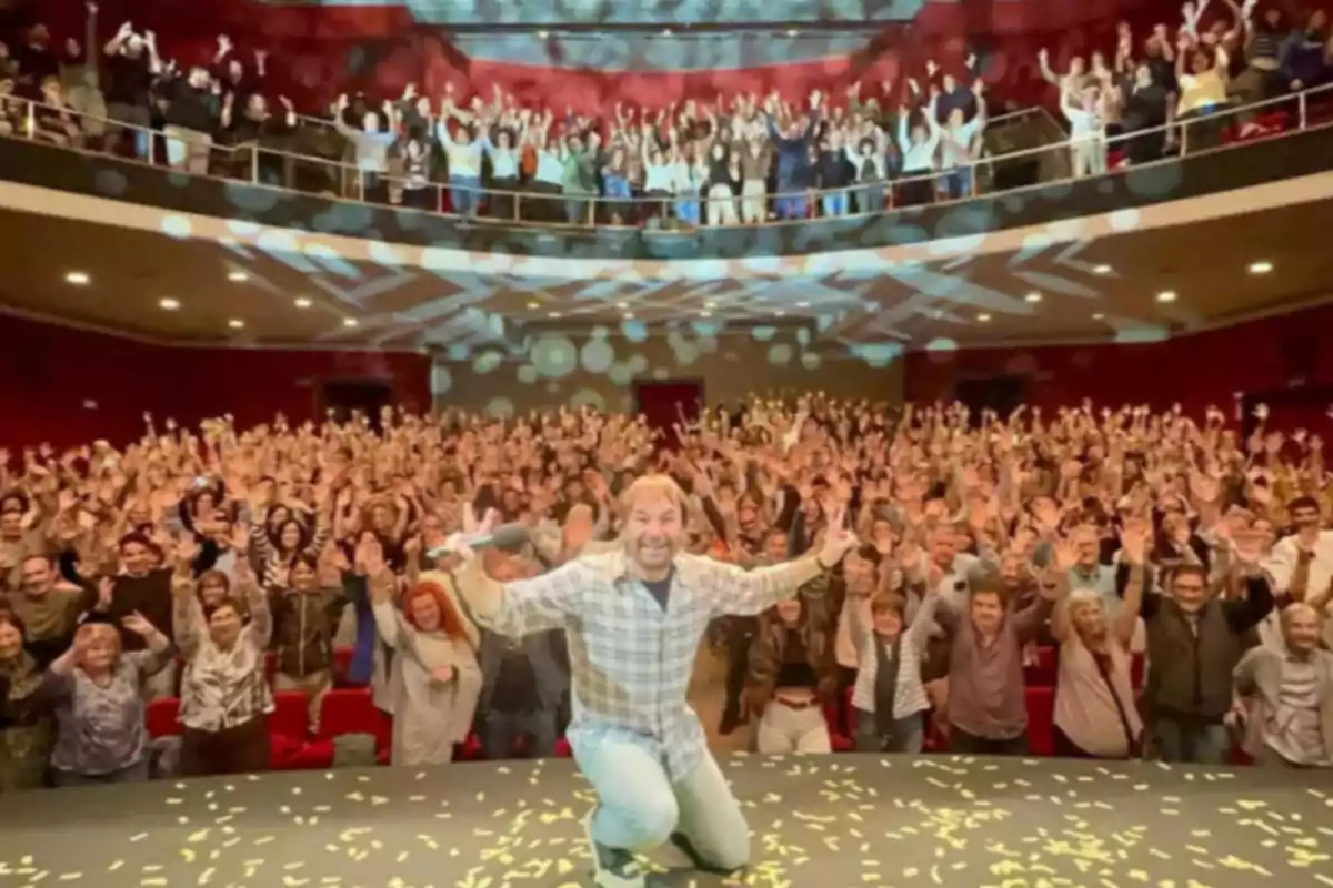 Un hombre sonriente arrodillado en el escenario de un teatro lleno de personas que levantan las manos celebrando juntos Un hombre sonriente arrodillado en el escenario de un teatro lleno de personas que levantan las manos celebrando juntos