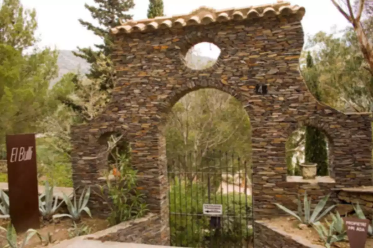 Entrada de pedra amb un portal de ferro envoltada de vegetació i cartells als costats Entrada de pedra amb un portal de ferro envoltada de vegetació i cartells als costats
