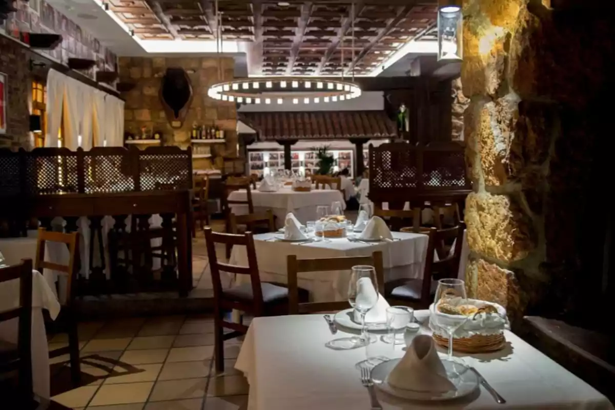 Restaurante Txistu elegante con mesas redondas cubiertas con manteles blancos, copas y servilletas dobladas, ambiente cálido con paredes de piedra y luz tenue Restaurante Txistu elegante con mesas redondas cubiertas con manteles blancos, copas y servilletas dobladas, ambiente cálido con paredes de piedra y luz tenue