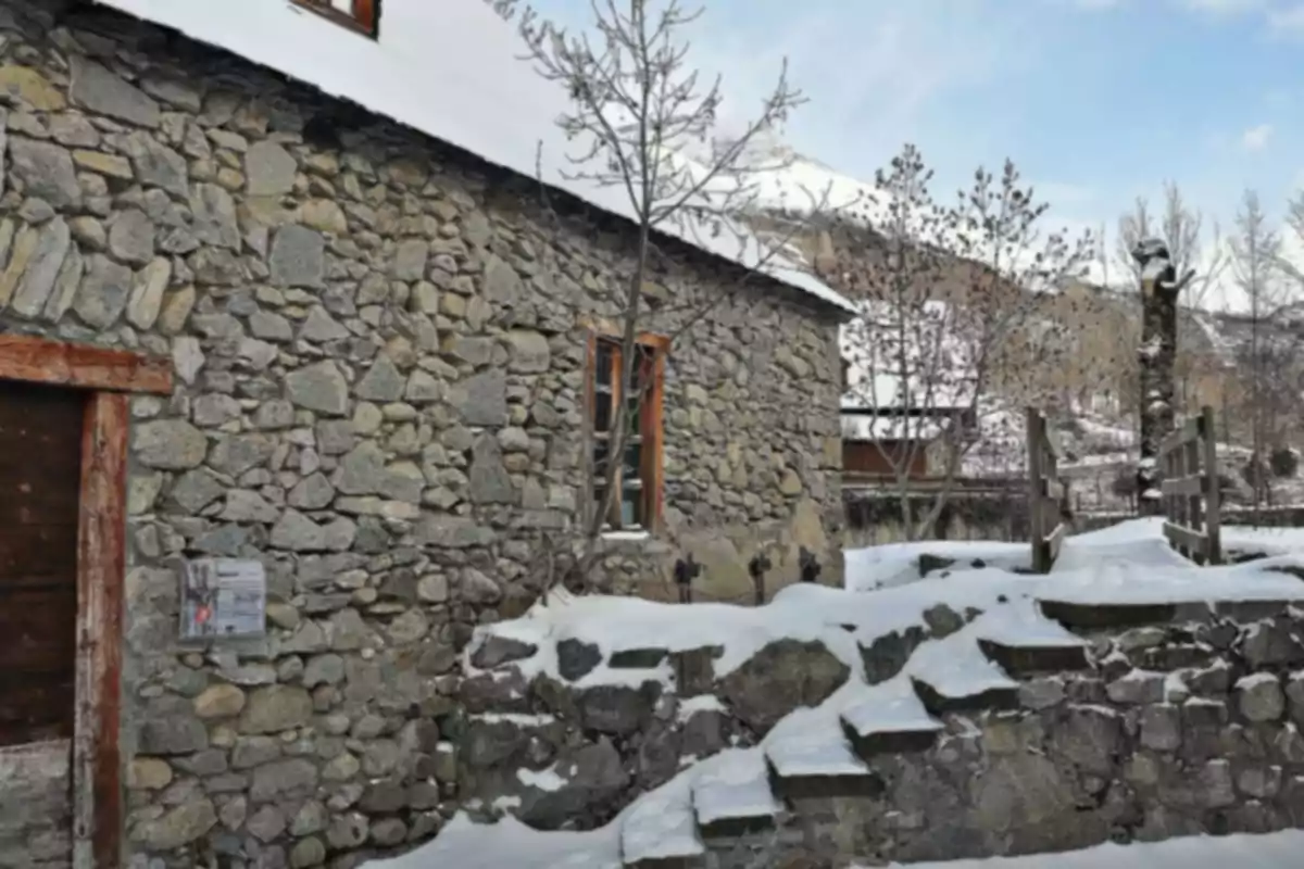 Casa de piedra con techo cubierto de nieve y escaleras exteriores rodeadas de árboles sin hojas en un entorno invernal Casa de piedra con techo cubierto de nieve y escaleras exteriores rodeadas de árboles sin hojas en un entorno invernal