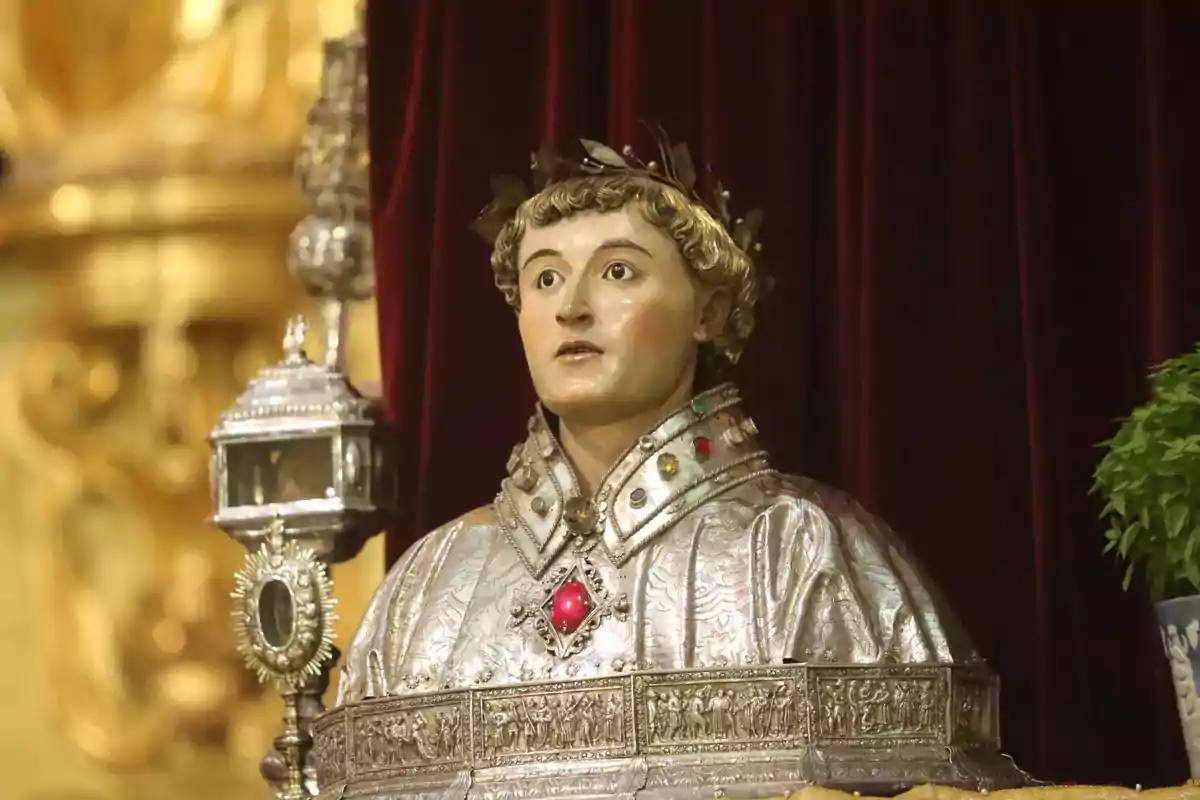 Busto religioso de aspecto antiguo hecho de metal decorado con piedras de colores y detalles ornamentales frente a un fondo de cortina roja Busto religioso de aspecto antiguo hecho de metal decorado con piedras de colores y detalles ornamentales frente a un fondo de cortina roja