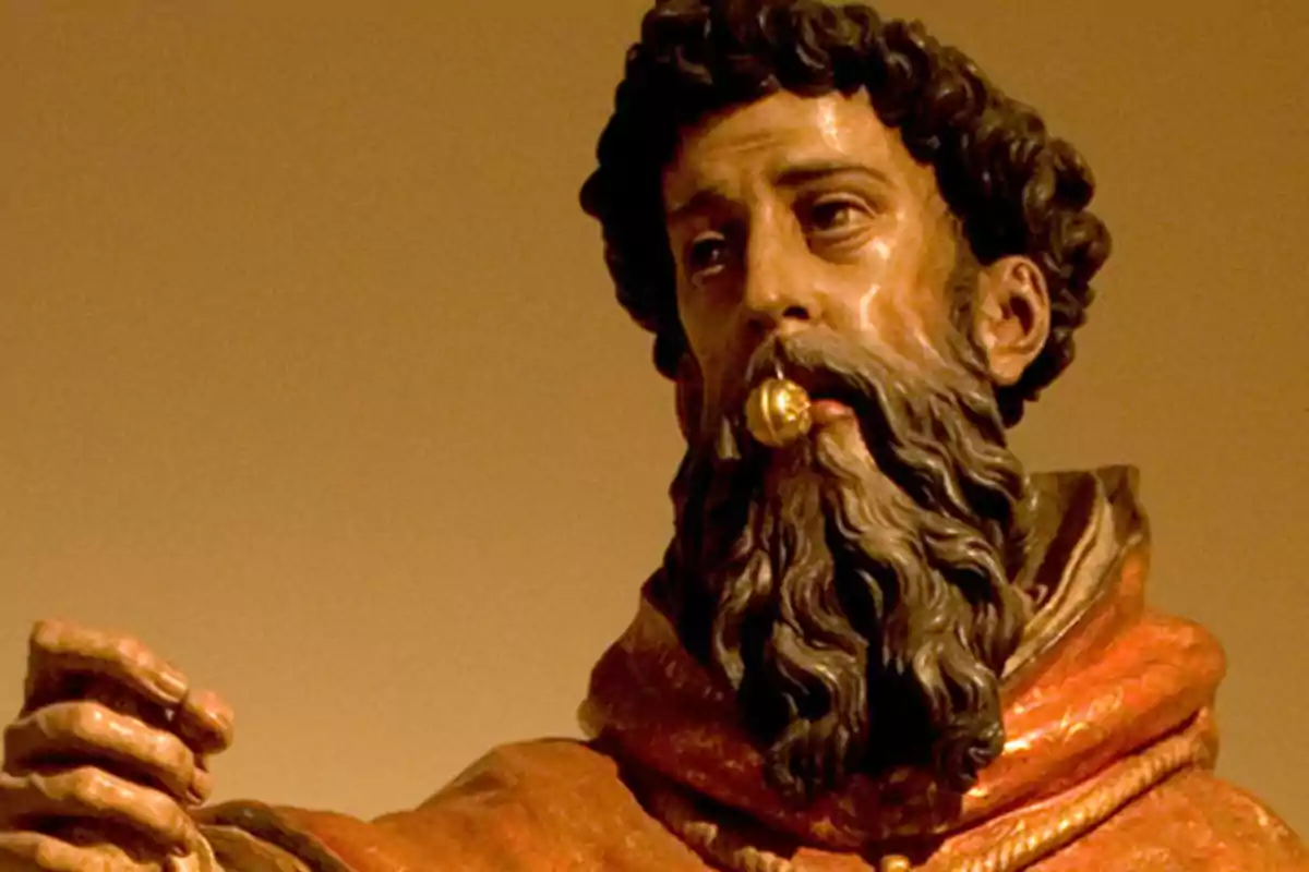 Escultura religiosa de un hombre barbudo con una bola dorada en la boca y túnica marrón rojiza sobre fondo marrón
