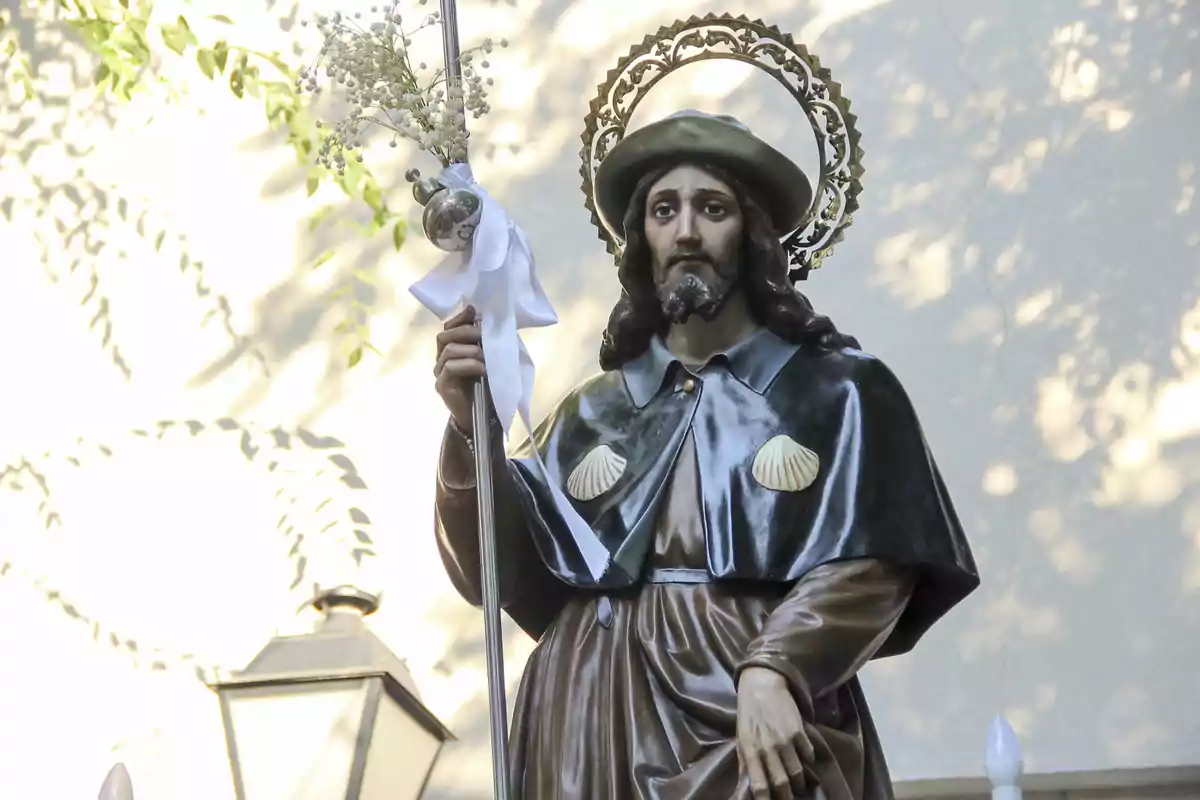Escultura religiosa de un hombre con sombrero y capa decorada con conchas sosteniendo un bastón adornado con flores y un lazo blanco Escultura religiosa de un hombre con sombrero y capa decorada con conchas sosteniendo un bastón adornado con flores y un lazo blanco