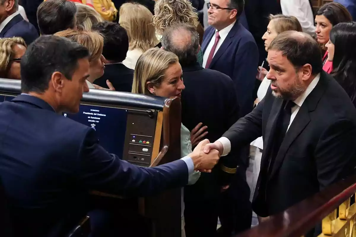 Dos hombres de traje se dan la mano en un entorno parlamentario rodeados de otras personas que conversan entre sí