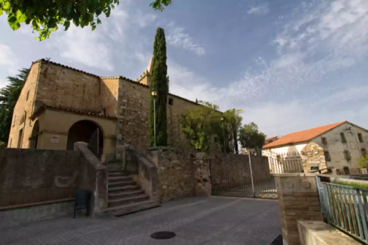 Edificio antiguo de piedra con escaleras y un ciprés en la entrada bajo un cielo parcialmente nublado Edificio antiguo de piedra con escaleras y un ciprés en la entrada bajo un cielo parcialmente nublado