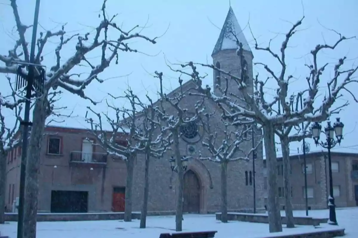 Església de pedra envoltada d’arbres sense fulles i coberts de neu en una plaça durant l’hivern Església de pedra envoltada d’arbres sense fulles i coberts de neu en una plaça durant l’hivern
