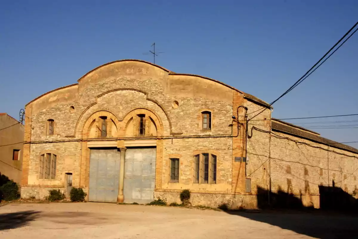 Façana d’una nau industrial antiga de pedra amb grans portes metàl·liques i finestres petites sota un cel serè Façana d’una nau industrial antiga de pedra amb grans portes metàl·liques i finestres petites sota un cel serè