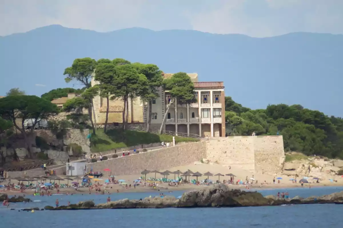 Platja concorreguda amb para-sols i banyistes davant d’un edifici antic envoltat d’arbres i muntanyes al fons Platja concorreguda amb para-sols i banyistes davant d’un edifici antic envoltat d’arbres i muntanyes al fons