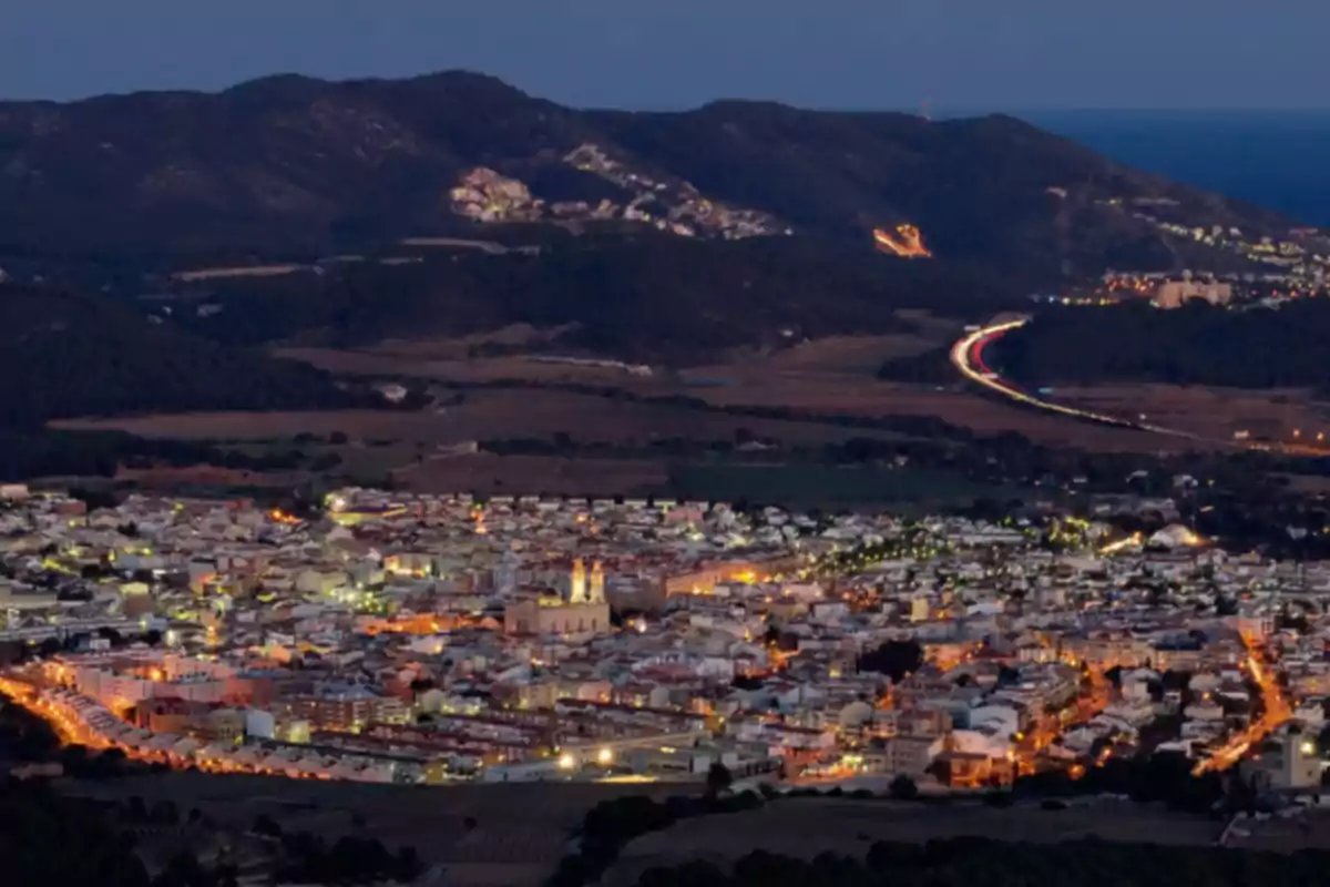 Vista panorámica de una ciudad iluminada al anochecer con montañas al fondo y una carretera con luces de autos Vista panorámica de una ciudad iluminada al anochecer con montañas al fondo y una carretera con luces de autos