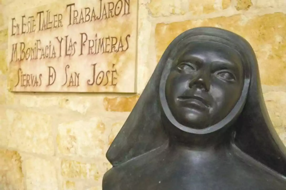 Busto de una mujer con hábito religioso de color oscuro frente a una pared de piedra y una placa conmemorativa en letras grandes. Busto de una mujer con hábito religioso de color oscuro frente a una pared de piedra y una placa conmemorativa en letras grandes.