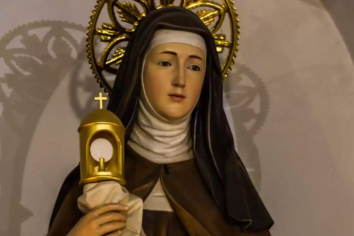 Escultura religiosa d’una dona amb hàbit fosc i aurèola daurada sostenint un objecte daurat amb una creu