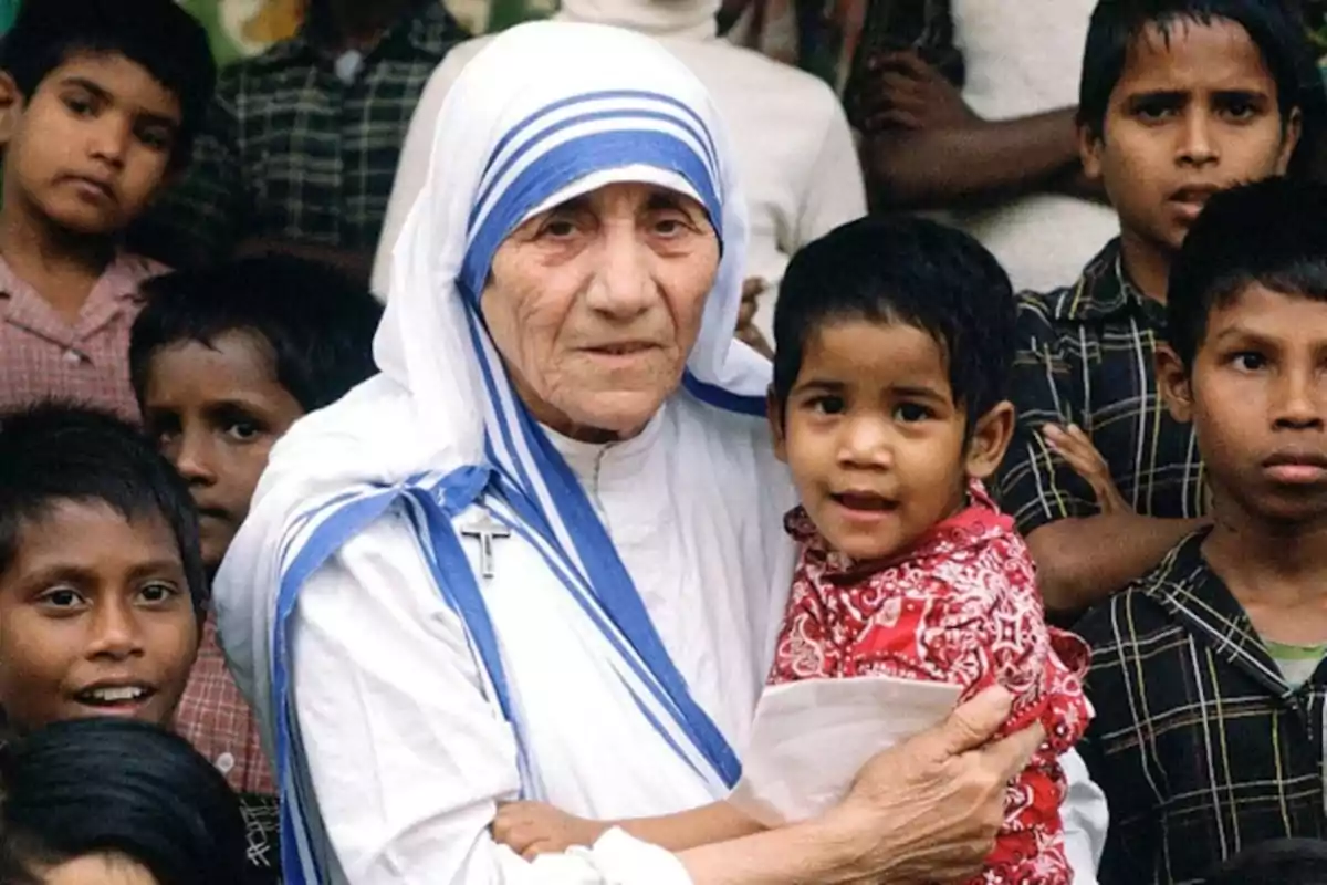 Una dona gran vestida amb un hàbit blanc i blau sosté en braços un nen petit envoltada de diversos nens que l'observen. Una dona gran vestida amb un hàbit blanc i blau sosté en braços un nen petit envoltada de diversos nens que l'observen.