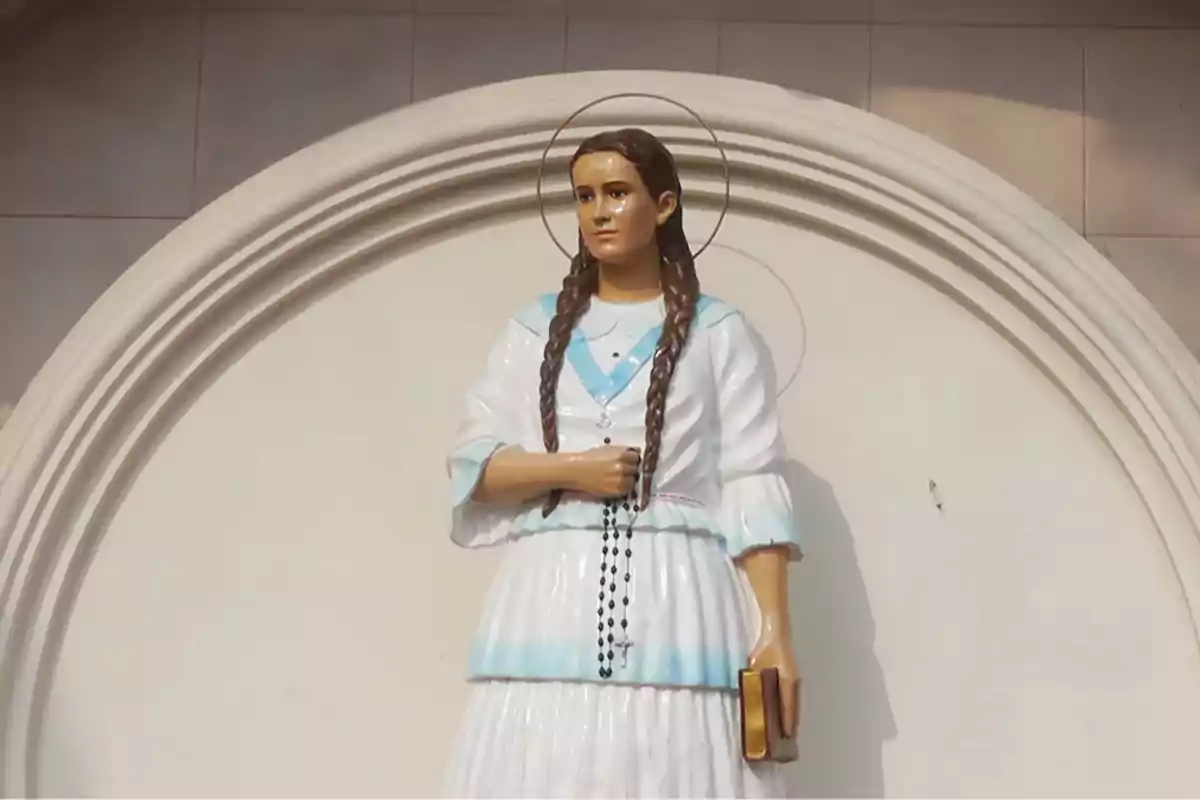 Escultura d’una jove amb trenes vestida de blanc i blau sostenint un rosari i un llibre amb una aurèola darrere del cap Escultura d’una jove amb trenes vestida de blanc i blau sostenint un rosari i un llibre amb una aurèola darrere del cap
