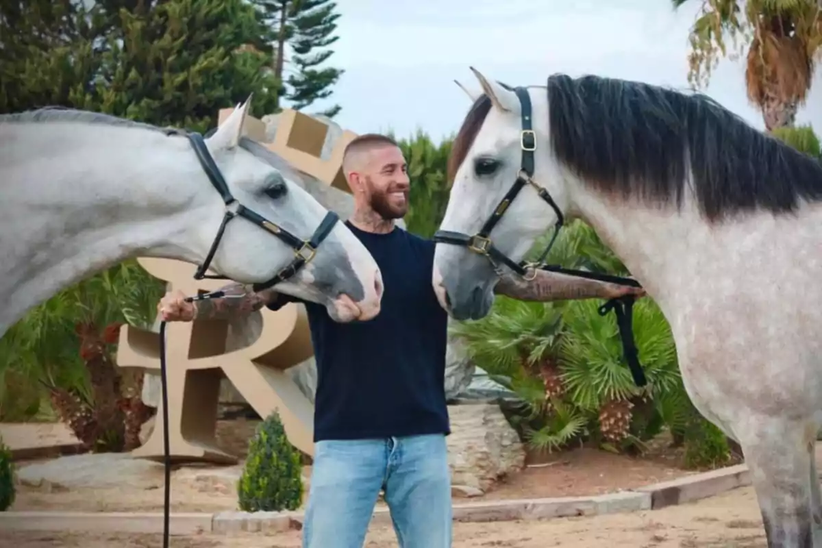 Sergio Ramos sonriente con barba y tatuajes sostiene a Yucatán de Ramos y Greco de Ramos II mientras los mira en un entorno al aire libre con palmeras y vegetación Sergio Ramos sonriente con barba y tatuajes sostiene a Yucatán de Ramos y Greco de Ramos II mientras los mira en un entorno al aire libre con palmeras y vegetación