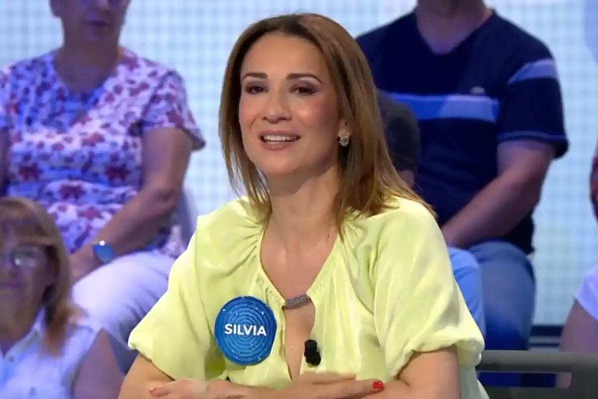 Silvia Jato sonriente con cabello castaño y blusa amarilla sentada en un Pasapalabra con una placa azul que dice Silvia y personas de fondo Silvia Jato sonriente con cabello castaño y blusa amarilla sentada en un Pasapalabra con una placa azul que dice Silvia y personas de fondo