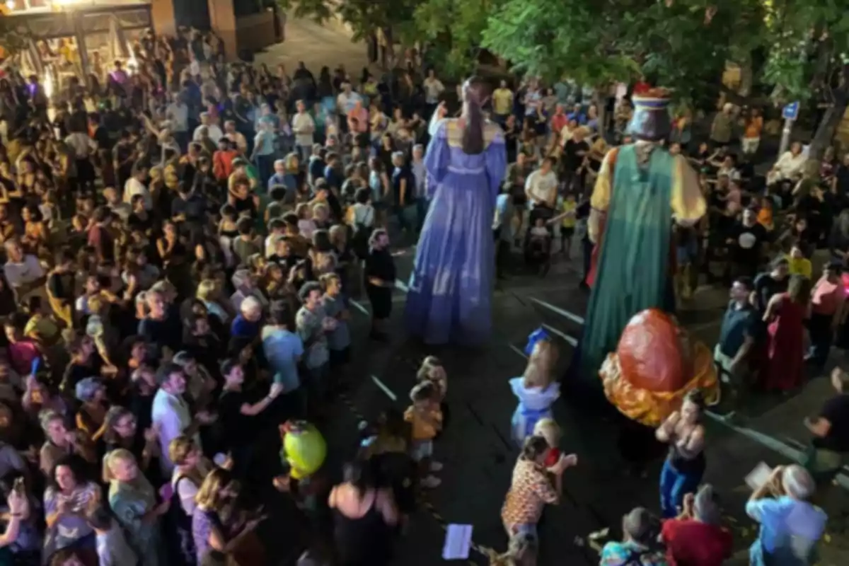 Multitud de persones reunides en una plaça durant la nit observant dues figures gegants vestides amb vestits acolorits. Multitud de persones reunides en una plaça durant la nit observant dues figures gegants vestides amb vestits acolorits.