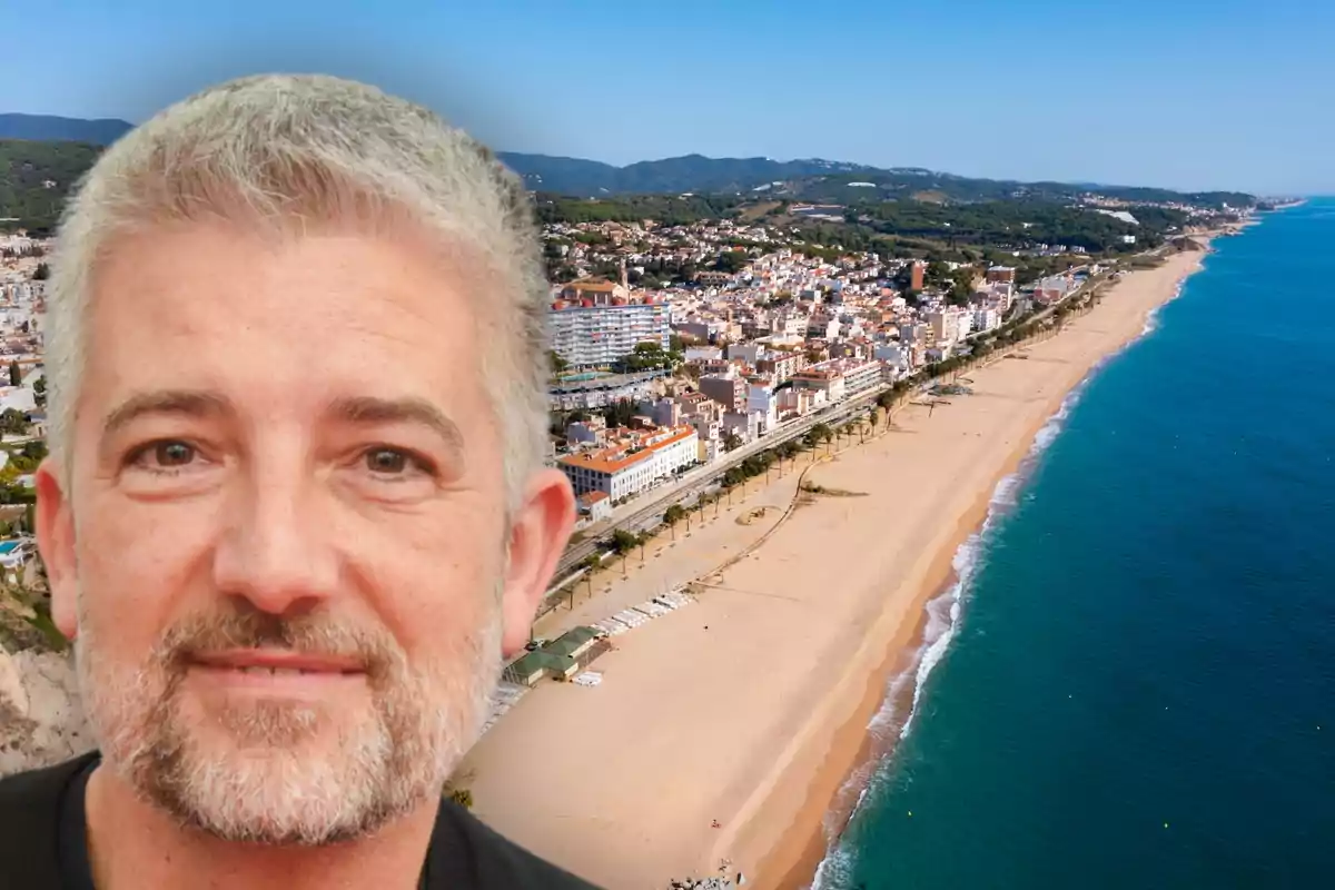 Hombre de cabello canoso y barba sonriente en primer plano con una playa y una ciudad costera al fondo