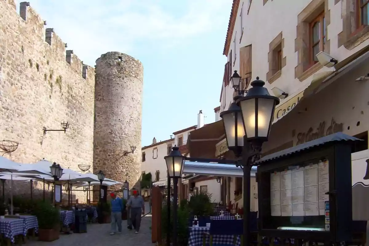 Carrer empedrat amb taules de restaurant a l’aire lliure al costat d’una muralla de pedra i una torre medieval Carrer empedrat amb taules de restaurant a l’aire lliure al costat d’una muralla de pedra i una torre medieval