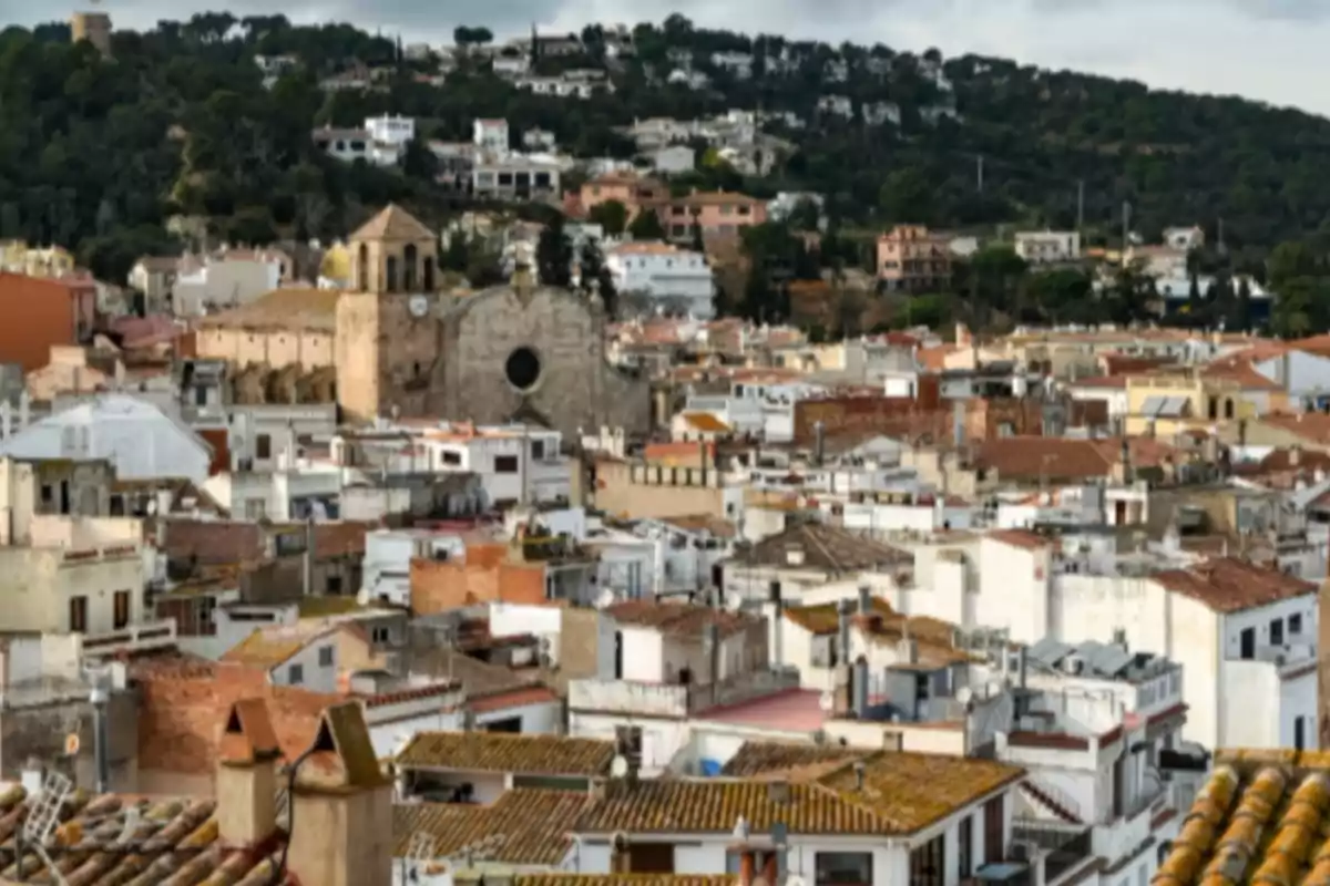 Vista panoràmica d’un poble amb cases de teulades vermelloses i una església antiga al fons envoltada de turons verds Vista panoràmica d’un poble amb cases de teulades vermelloses i una església antiga al fons envoltada de turons verds