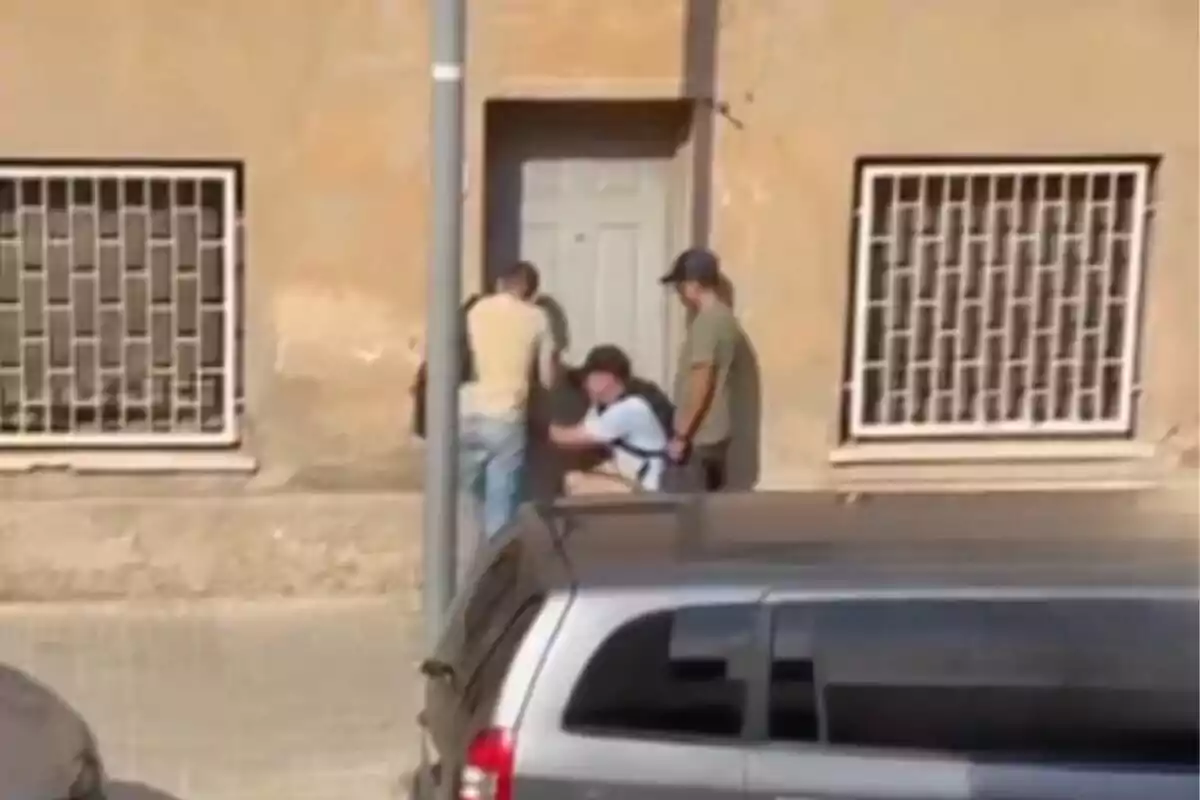 Tres personas están frente a una puerta en la entrada de un edificio, una de ellas parece estar manipulando la cerradura mientras las otras observan y hay una camioneta gris estacionada en primer plano.