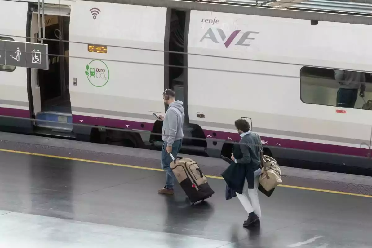 Dos personas caminan por el andén de una estación de tren junto a un tren AVE de Renfe llevando maletas y bolsos. Dos personas caminan por el andén de una estación de tren junto a un tren AVE de Renfe llevando maletas y bolsos.