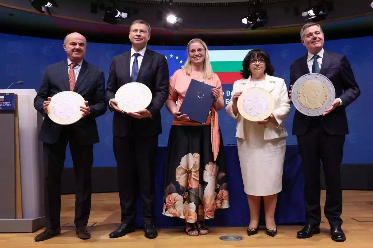 Cinc persones vestides de manera formal posen en un escenari sostenint grans rèpliques de monedes davant d’un fons blau amb la bandera de Bulgària i la bandera de la Unió Europea. Cinc persones vestides de manera formal posen en un escenari sostenint grans rèpliques de monedes davant d’un fons blau amb la bandera de Bulgària i la bandera de la Unió Europea.