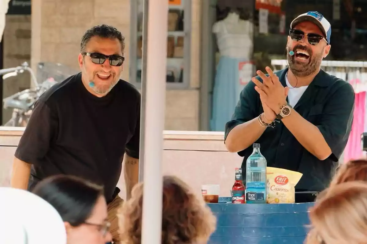 Dos hombres sonríen y se divierten mientras hablan con micrófonos frente a una mesa con botellas y una bolsa de papas, rodeados de público en un evento al aire libre Dos hombres sonríen y se divierten mientras hablan con micrófonos frente a una mesa con botellas y una bolsa de papas, rodeados de público en un evento al aire libre