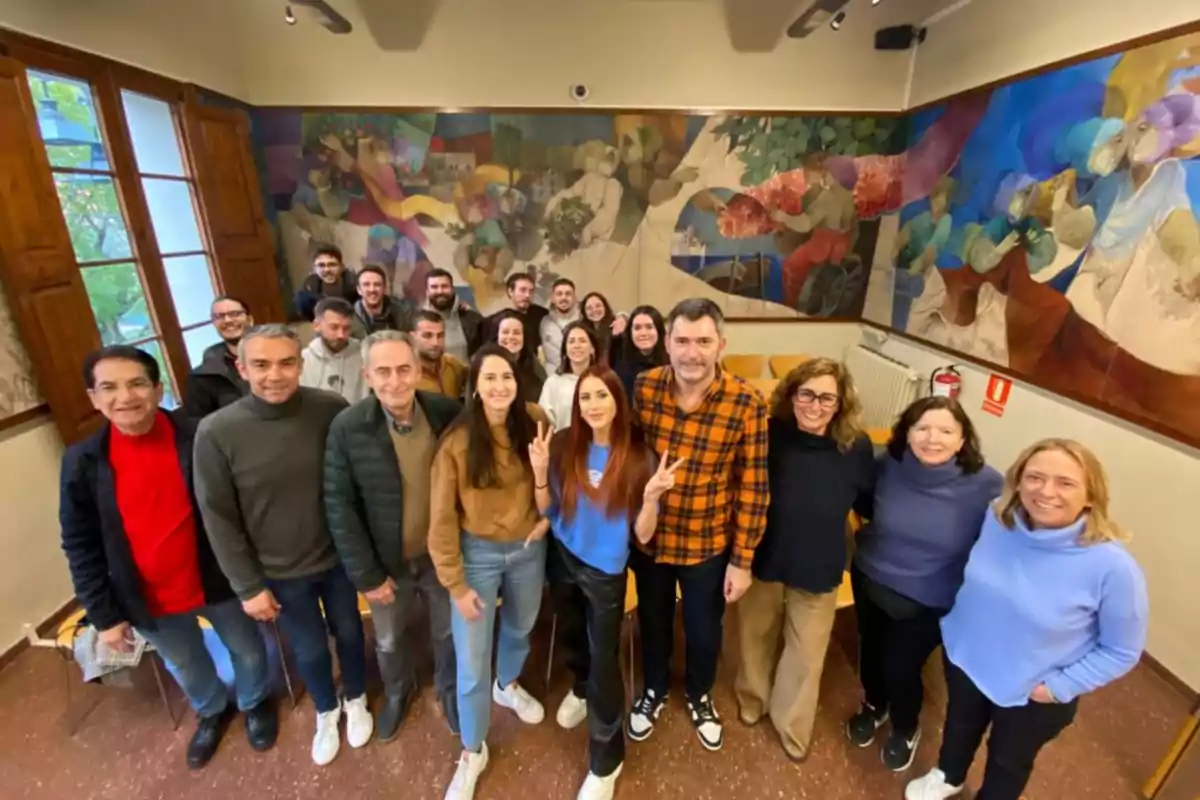 Grup de persones posant i somrient dins d’una sala amb murals colorits a les parets i finestres de fusta obertes al fons Grup de persones posant i somrient dins d’una sala amb murals colorits a les parets i finestres de fusta obertes al fons