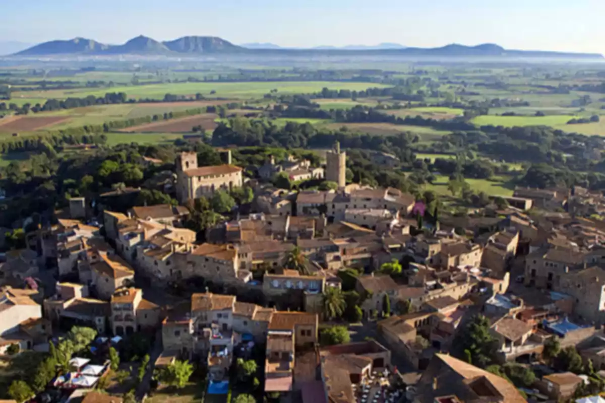Vista aérea de un pueblo medieval rodeado de campos verdes y montañas al fondo Vista aérea de un pueblo medieval rodeado de campos verdes y montañas al fondo