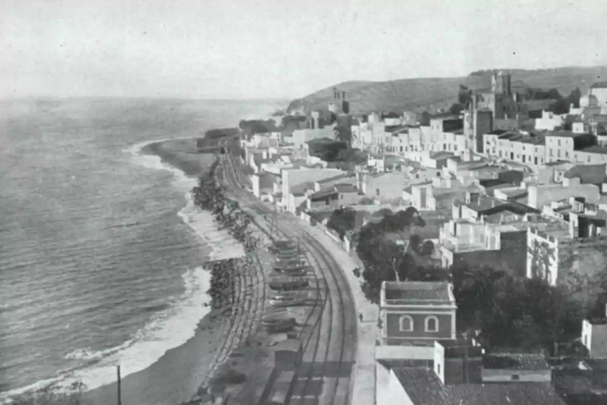 Vista antigua en blanco y negro de un pueblo costero con vías de tren junto al mar y casas alineadas cerca de la playa Vista antigua en blanco y negro de un pueblo costero con vías de tren junto al mar y casas alineadas cerca de la playa