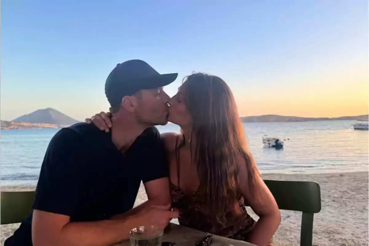 Xavi Alonso y Nagore Aranburu besándose sentada en una mesa frente al mar al atardecer con un paisaje de montañas y barcos al fondo Xavi Alonso y Nagore Aranburu besándose sentada en una mesa frente al mar al atardecer con un paisaje de montañas y barcos al fondo