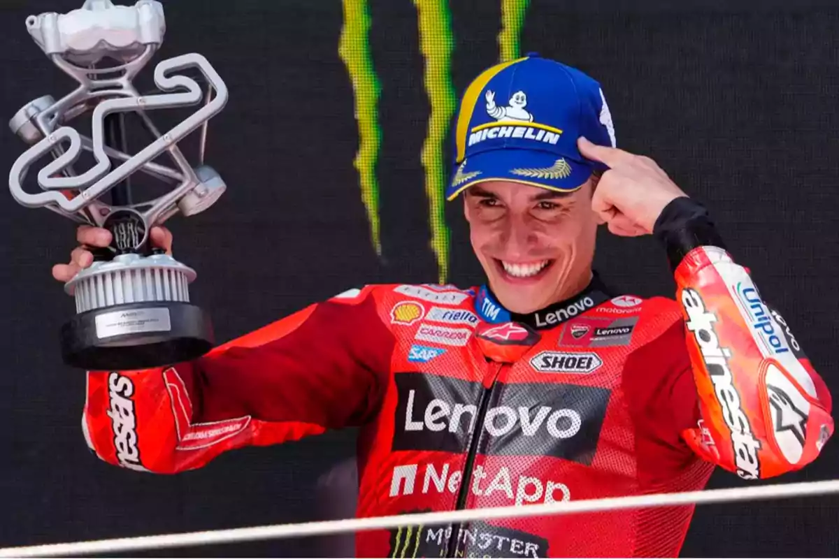 Marc Márquez, a smiling motorcycle racer in a red Ducati suit celebrating on the podium while holding a trophy and pointing to his blue Michelin cap Marc Márquez, a smiling motorcycle racer in a red Ducati suit celebrating on the podium while holding a trophy and pointing to his blue Michelin cap