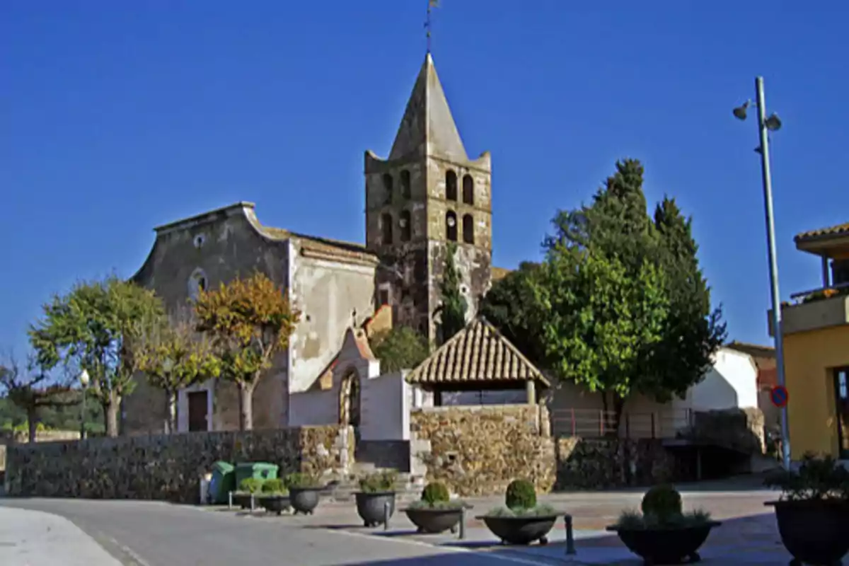 Església antiga amb campanar de pedra envoltada d’arbres i testos en una plaça sota un cel serè Església antiga amb campanar de pedra envoltada d’arbres i testos en una plaça sota un cel serè
