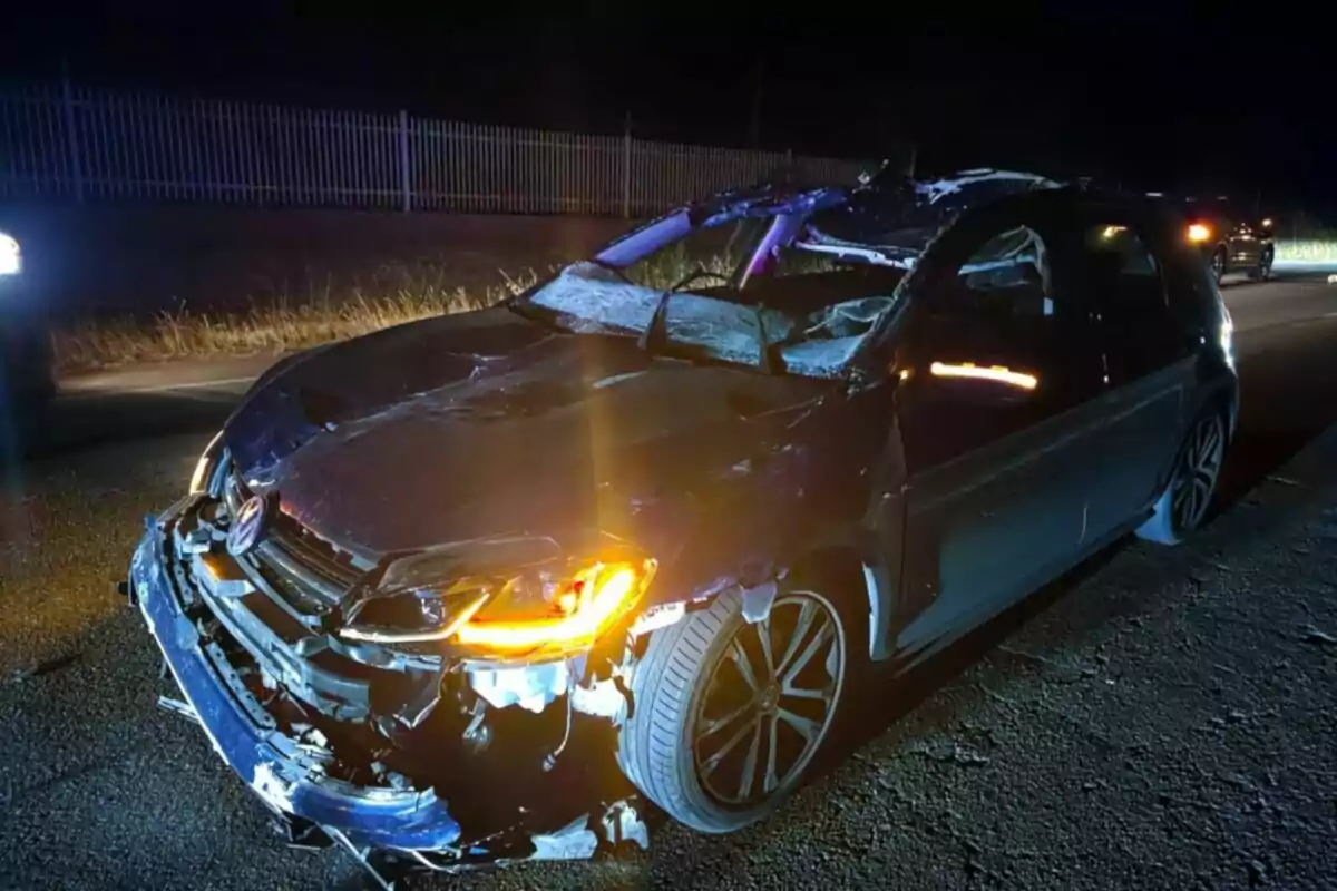 Auto negro con graves daños en la carrocería y el parabrisas, estacionado en una carretera durante la noche con las luces encendidas.