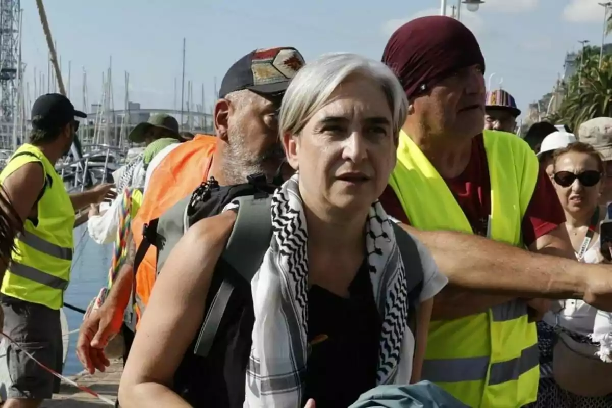 Mujer de cabello canoso con pañuelo blanco y negro al cuello rodeada de personas con chalecos reflectantes en un puerto durante el día